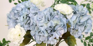 En närbild på en bukett med konstgjorda ljusblå hortensior med fylliga blomhuvuden, blandade med vita nejlika blommor och gröna blad.