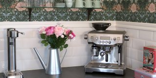 Ett stilrent kök med mörkgrön tapet med blommigt mönster och vitt kakel. På köksbänken står en bukett med rosa rosor i en silverfärgad vattenkanna, en sodastreamer i stål och en espressomaskin. En hylla ovanför rymmer kaffemuggar.